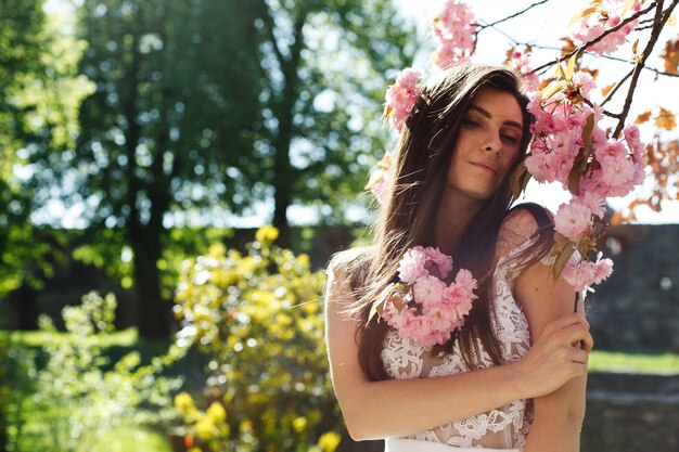 Encantadora mujer joven en vestido rosa posa ante un árbol de sakura lleno de flores rosadas