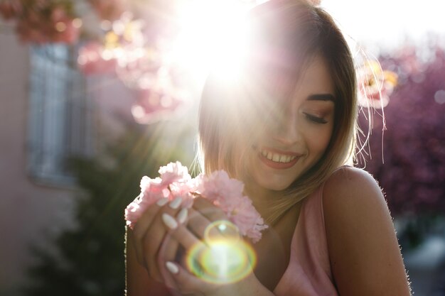 Encantadora mujer joven en vestido rosa posa ante un árbol de sakura lleno de flores rosadas