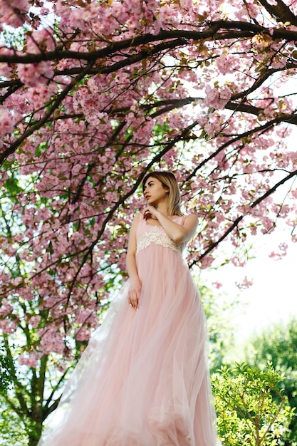 Encantadora mujer joven en vestido rosa posa ante un árbol de sakura lleno de flores rosadas