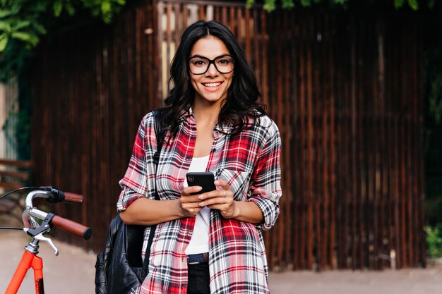 Encantadora mujer latina con sonrisa feliz con smartphone en la calle. Hermosa chica europea en traje casual de pie junto a la bicicleta.