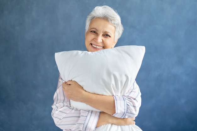 Encantadora mujer madura alegre en pijama de rayas con aspecto feliz porque durmió lo suficiente, abrazando la almohada de plumas blancas y sonriendo ampliamente