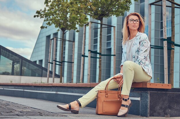 Encantadora mujer de moda con ropa elegante y gafas con un bolso, sentada en un banco contra un rascacielos.