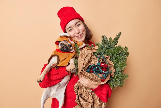 Encantadora mujer morena complacida posa con un perro pug y atributos de vacaciones se prepara para las celebraciones de Navidad o Año Nuevo viste un suéter rojo y un sombrero disfruta del tiempo festivo con poses de mascotas favoritas en el interior