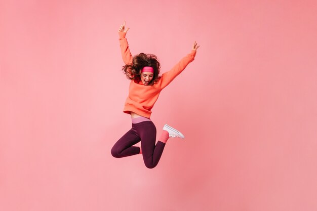 Encantadora mujer morena en diadema deportiva rosa y chándal salta en pared rosa aislada