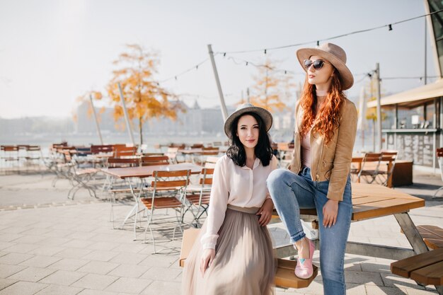 Encantadora mujer morena en falda larga sentada en un café al aire libre con una amiga en elegante sombrero