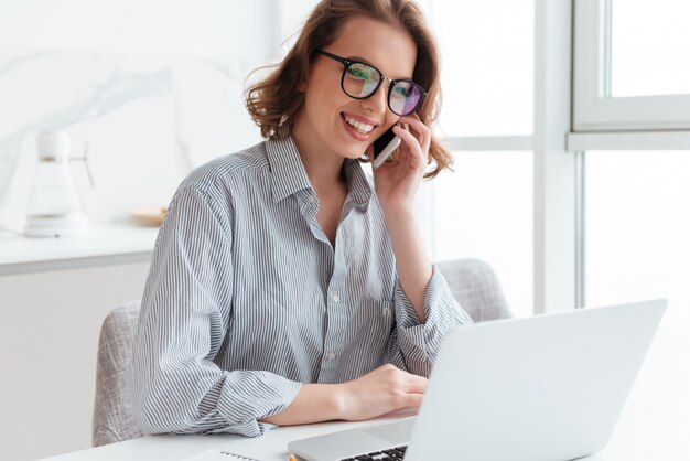 Encantadora mujer morena con gafas y ropa casual hablando por teléfono móvil mientras está sentado en la mesa y mirando la pantalla del portátil