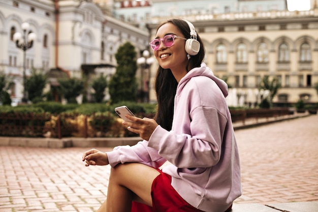Foto gratuita encantadora mujer morena con sudadera con capucha rosa y gafas de sol mira hacia otro lado, sostiene el teléfono y escucha música en auriculares afuera