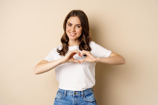 Encantadora mujer mostrando corazón, signo de amor y sonriendo, de pie sobre fondo beige en camiseta blanca, fondo beige.