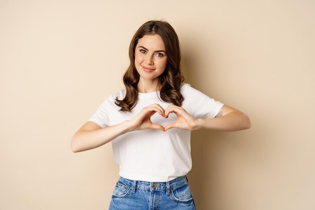 Encantadora mujer mostrando signo de amor de corazón y sonriendo de pie sobre fondo beige en camiseta blanca beig...