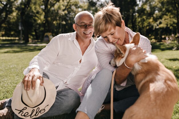 Foto gratuita encantadora mujer con peinado corto sonriendo, jugando con corgi y sentada sobre la hierba con el hombre de pelo gris con sombrero en camisa blanca al aire libre.