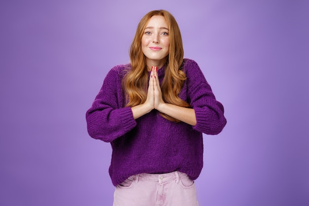 Encantadora mujer pelirroja linda con cabello largo y pecas en un suéter púrpura tomados de la mano en oración haciendo una sonrisa de esperanza y un ángel que pide ayuda o misericordia, pidiendo con súplica.