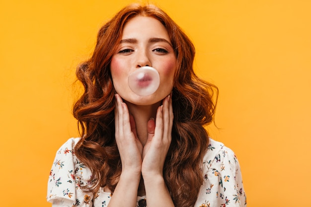 Encantadora mujer pelirroja rizada en top blanco hace burbuja de chicle. Retrato de mujer joven sobre fondo naranja.