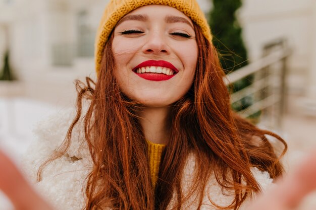 Encantadora mujer de pelo largo con sombrero riendo con los ojos cerrados. Foto al aire libre de una entusiasta chica de jengibre que expresa felicidad en invierno.