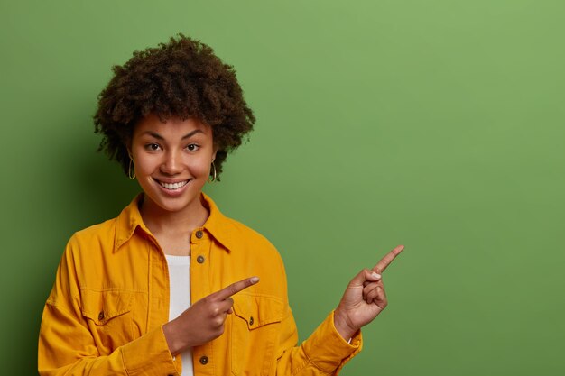 Encantadora mujer de piel oscura con cabello afro señala con el dedo índice a la derecha, sugiere ir en esta dirección, demuestra un producto increíble, usa una chaqueta amarilla,