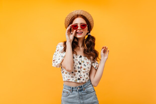 Encantadora mujer rizada en elegante blusa recortada y falda de mezclilla lleva gafas de sol rojas. Mujer en navegante mira a cámara sobre fondo naranja.