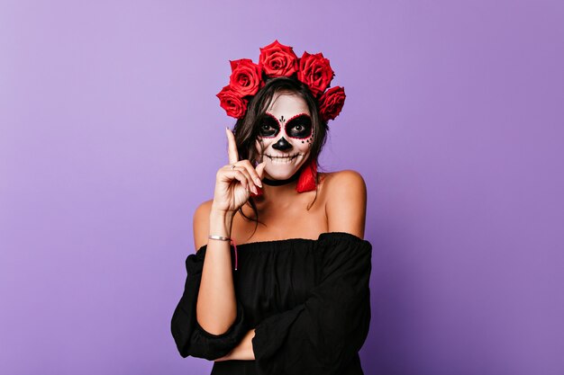 Encantadora mujer con rosas en el pelo negro esperando la fiesta de halloween. Feliz modelo de mujer latina con pintura de cara de vampiro sonriendo