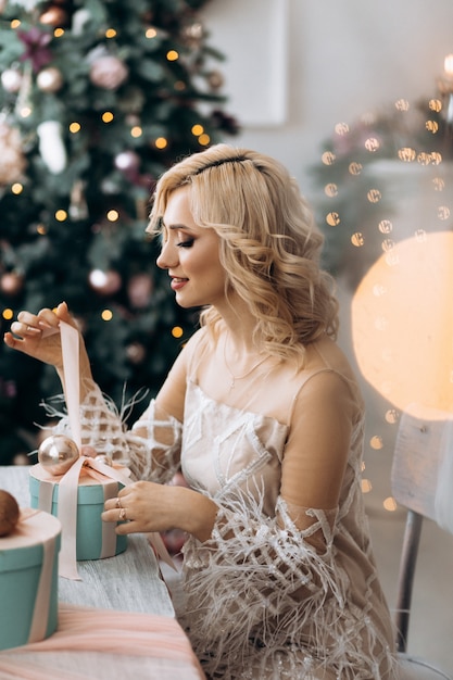 Encantadora mujer rubia abre cajas presentes sentadas ante un árbol de Navidad