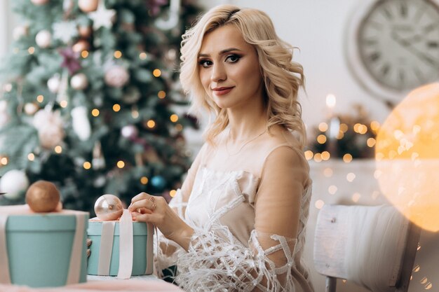 Encantadora mujer rubia abre cajas presentes sentadas ante un árbol de Navidad