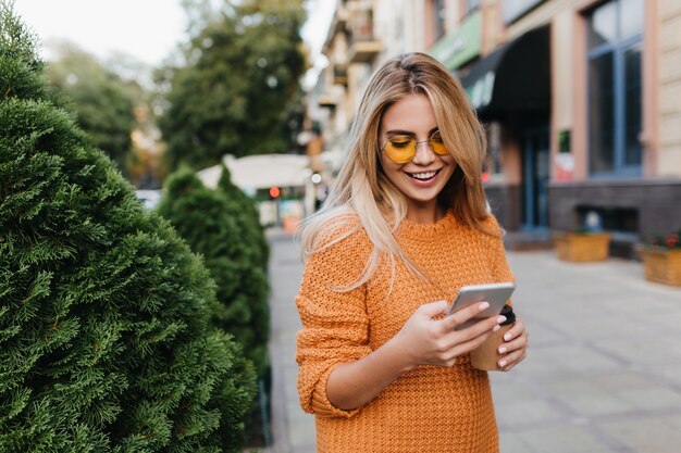 Encantadora mujer rubia caminando por arbustos verdes con sonrisa, llevando smartphone y taza de café