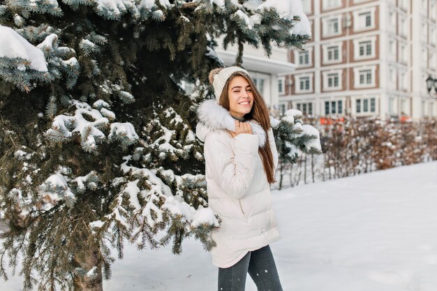 Encantadora mujer rubia con chaqueta blanca y jeans negros posando durante la caminata en el parque de invierno. Foto al aire libre de una mujer muy de moda que se divierte en la mañana de diciembre.