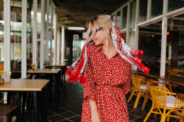 Encantadora mujer rubia feliz caminando afuera con un vestido rojo brillante y un chal en la cabeza