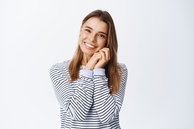 Encantadora mujer rubia mirando algo adorable o hermoso, suspirando romántico, tomados de la mano cerca de las mejillas y sonriendo, pared blanca