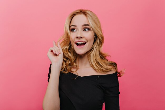 Encantadora mujer rubia posando con sonrisa de sorpresa. Foto interior de niña blanca interesada en vestido negro aislado en la pared rosa.