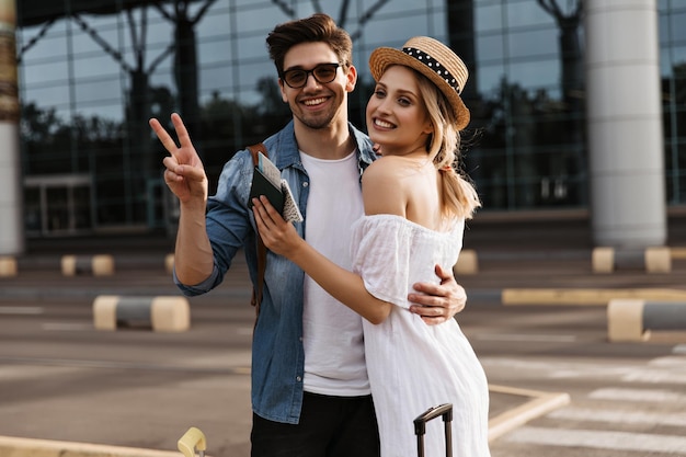 Encantadora mujer rubia con sombrero vestido blanco y alegre hombre moreno con gafas de sol, chaqueta de mezclilla, sonríe y muestra vsign