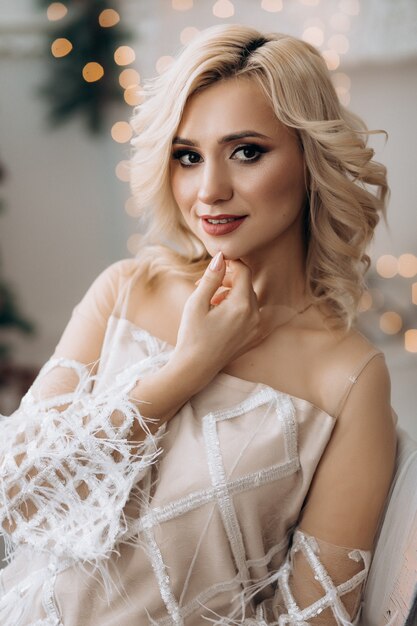 Encantadora mujer rubia con vestido blanco posa en una habitación con un gran árbol de Navidad