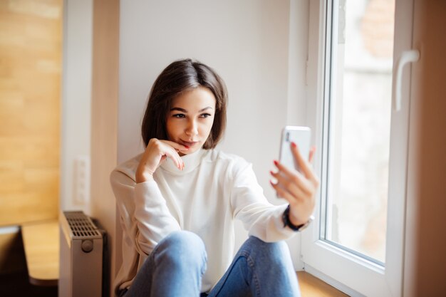 Encantadora mujer sentada en el alféizar de la ventana en blue jeans con teléfono haciendo selfie