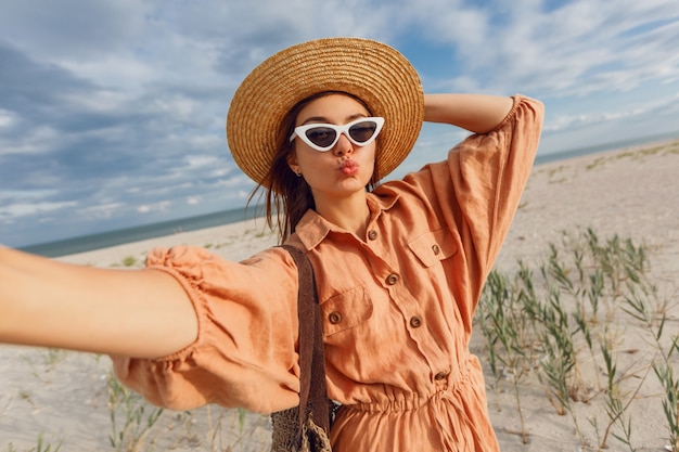 Encantadora mujer sonriente haciendo autorretrato y disfrutando de unas vacaciones cerca del océano. El uso de gafas de sol retro de moda y sombrero de paja.