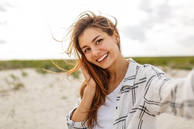 Encantadora mujer sonriente haciendo autorretrato y disfrutando de unas vacaciones cerca del océano.