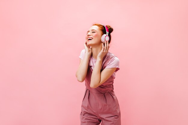 Encantadora mujer con sonrisa mira a cámara, sosteniendo el teléfono inteligente en sus manos. mujer en vestido rosa escuchando música en auriculares.