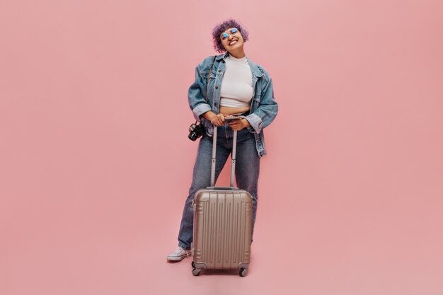 Encantadora mujer con sonrisa en su rostro con chaqueta de mezclilla de gran tamaño, pantalones ajustados y top blanco sostiene la maleta y la cámara en rosa.