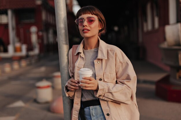 Encantadora mujer sosteniendo una taza de café en la ciudad Morena jovencita en chaqueta de mezclilla rosa gafas de sol y jeans con cinturón negro posando en la calle