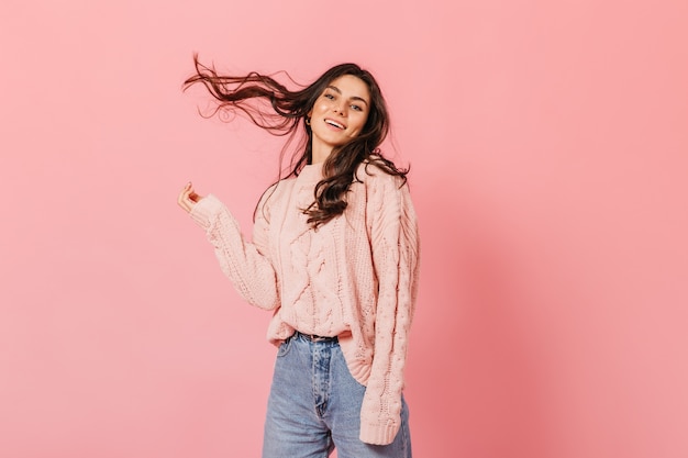 Encantadora mujer en suéter rosa juega con el pelo sobre fondo aislado. Lady in jeans se ríe y mira a la cámara.