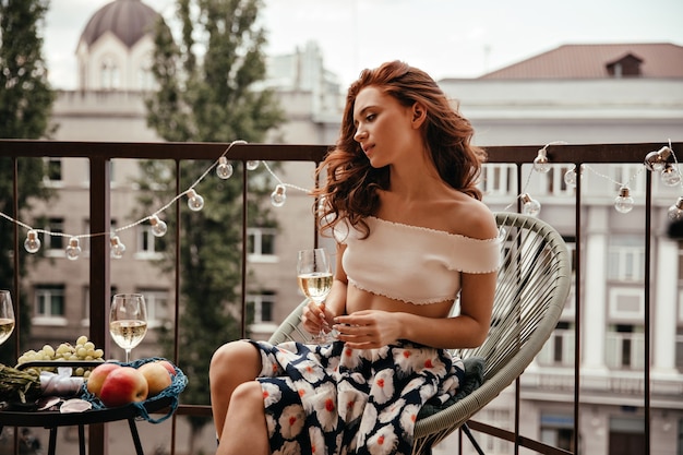 Encantadora mujer en traje floral sostiene copa de vino y descansa en la terraza