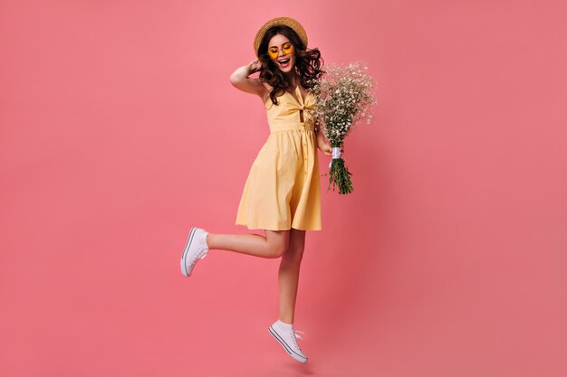 Encantadora mujer en vestido amarillo salta sobre pared rosa y sostiene ramo