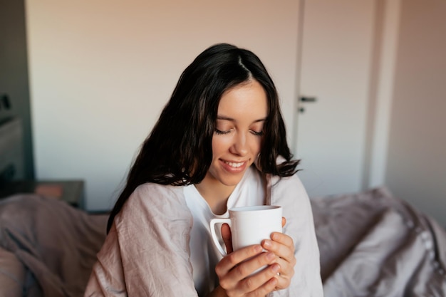 Encantadora niña adorable con cabello oscuro sostiene una taza de café y se cubre con una manta en el dormitorio por la mañana bajo el sol Concepto de hogar Despierta por la mañana en casa