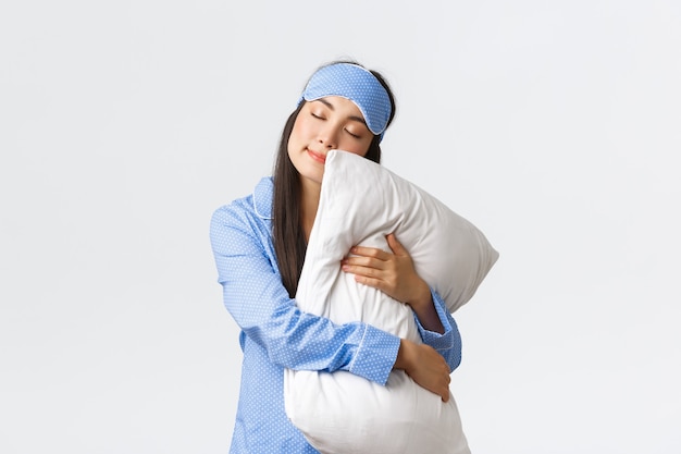 Encantadora niña asiática durmiendo en antifaz y pijama, abrazando la almohada y acostada con los ojos cerrados, soñando, durmiendo por la noche, posando sobre fondo blanco de ensueño. Copia espacio