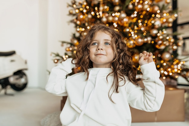 Encantadora niña bonita con rizos en ropa blanca sonriendo delante del árbol de Navidad con luces