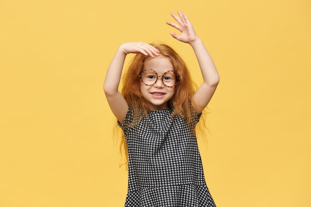 Encantadora niña caucásica pelirroja con vestido y anteojos redondos gesticulando emocionalmente, emocionado con buenas noticias positivas, con sonrisa feliz