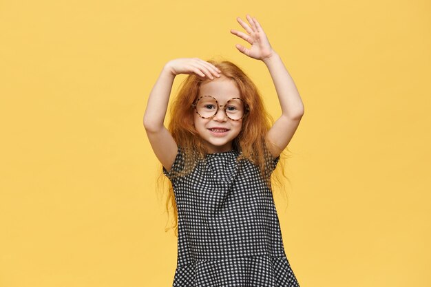 Encantadora niña caucásica pelirroja con vestido y anteojos redondos gesticulando emocionalmente, emocionado con buenas noticias positivas, con sonrisa feliz