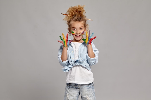 Foto gratuita encantadora niña con un cepillo en su elegante peinado, vestida con una camisa azul y una camiseta blanca. levantó sus manos pintadas, mirando a la cámara y sonriendo ampliamente, sobre un fondo gris.