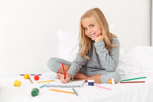 Encantadora niña dibujando con lápices sobre cama blanca