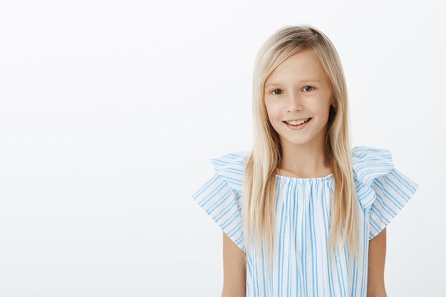 Encantadora niña feliz presentándose a sus nuevos compañeros. Retrato de encantadora hija adorable despreocupada con cabello rubio en blusa azul, de pie casualmente sobre la pared gris y sonriente