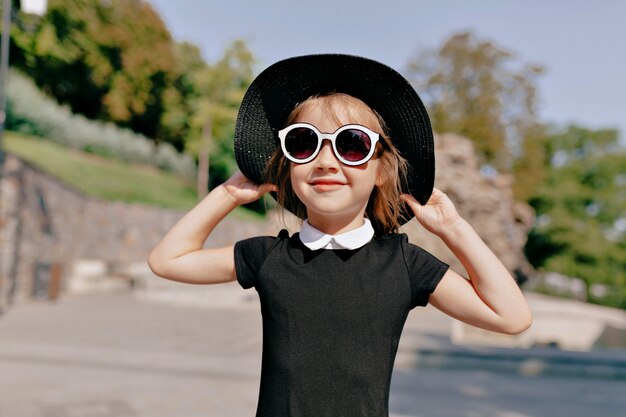 Encantadora niña linda con sombrero y gafas de sol caminando en el parque en un día cálido y soleado