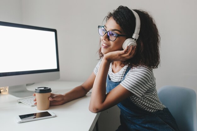 Encantadora niña mulata sentada en la mesa con computadora y teléfono inteligente y apuntando la cara con la mano