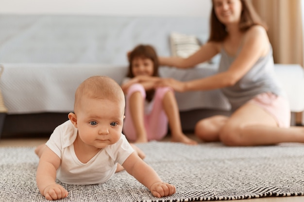 Encantadora niña pequeña arrastrándose por el suelo sobre la alfombra en la sala de estar, niña jugando en casa con la madre y la hermana en el fondo, infancia feliz.