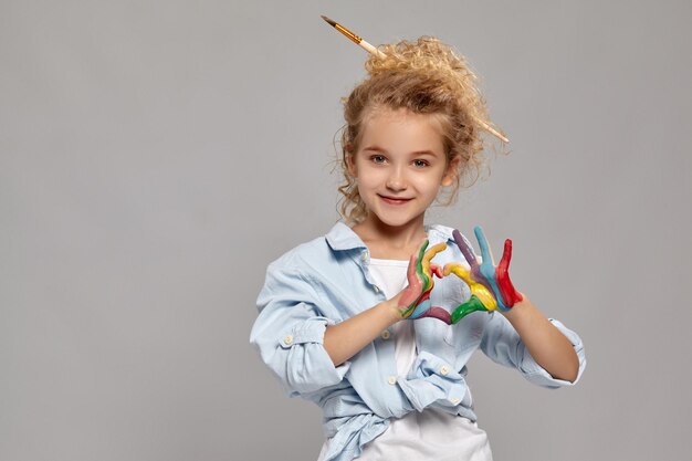 Encantadora niña pequeña con un cepillo en su elegante cabello rubio rizado, vestida con una camisa azul y una camiseta blanca. Ha doblado sus dedos pintados en forma de corazón y sonriendo, sobre un fondo gris.